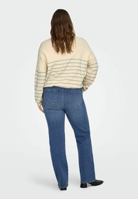 Femme aux longs cheveux bruns portant un pull crème à fines rayures bleues, un jean bleu coupe droite et des chaussures noires à talons blocs, vue de dos.