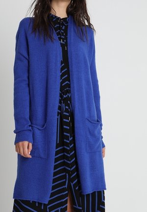 Femme portant un long cardigan bleu ouvert devant une robe noire avec des rayures géométriques bleues, debout devant un fond clair uni.