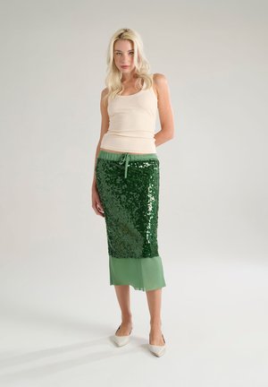Jupe verte en sequins avec un ourlet inférieur transparent, associée à un débardeur beige. La jupe possède une taille à cordon et une silhouette ajustée.