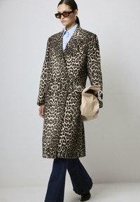 Femme portant un long manteau en imprimé léopard, une chemise bleue, un jean flare noir, des lunettes de soleil, tenant un clutch beige plié devant un fond uni.