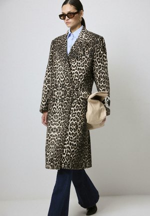 Femme portant un long manteau en imprimé léopard, une chemise bleue, un jean flare noir, des lunettes de soleil, tenant un clutch beige plié devant un fond uni.