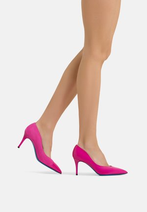 Gekreuzte Beine einer Frau, die leuchtend pinke spitze High Heels trägt, vor weißem Hintergrund.