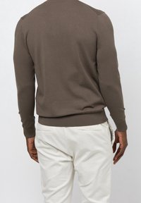 Man i brun långärmad tröja och ljusbeiga byxor, stående med ryggen mot kameran framför en enfärgad bakgrund.