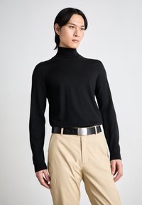 Black turtleneck sweater, ribbed cuffs, fitted design, paired with beige trousers and a black belt featuring a silver buckle.