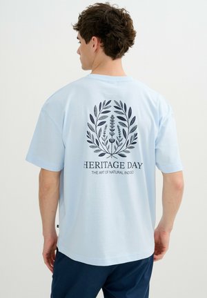 Hombre de espaldas que lleva una camiseta azul claro con diseño de corona botánica y texto "DÍA DEL PATRIMONIO EL ARTE DEL ÍNDIGO NATURAL" en la espalda.