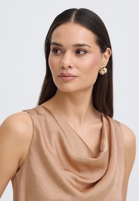 Femme aux longs cheveux foncés portant des boucles d'oreilles florales dorées et un haut en satin beige sans manches, regardant légèrement vers sa droite sur un fond blanc.