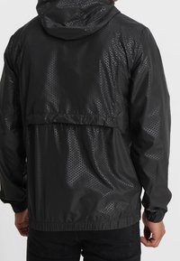 Veste noire avec capuche, motif hexagonal texturé, poignets et ourlet élastiques, matériau léger et ouverture arrière pour la respirabilité.