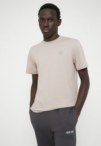 Νεαρός άνδρας με τα χέρια στις τσέπες σκούρων γκρι παντελονιών, φορώντας μια μπεζ μπλούζα με μικρό λογότυπο στο στήθος, μπροστά σε λευκό φόντο.