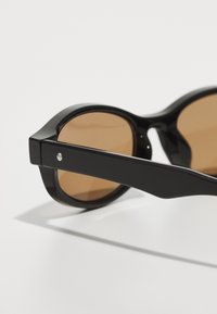 Black sunglasses with rounded frames, brown lenses, and sleek plastic material. Features a metallic accent on the arms.