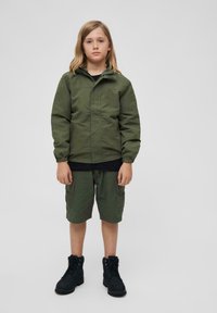 Veste vert olive avec une fermeture éclair à l'avant, poignets élastiques ; t-shirt noir en dessous. Shorts olive avec poches et bottes noires. Design simple.