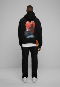 Felpa nera con un grafico colorato sul retro, abbinata a pantaloni cargo neri e sneakers bianche. Presenta un'etichetta sul lato.