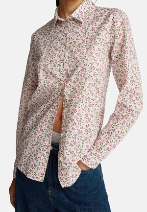 Hvid skjorte med lange ærmer og lille pink og grøn blomsterprint, iført delvist åben med den ene hånd i lommen på blå jeans.