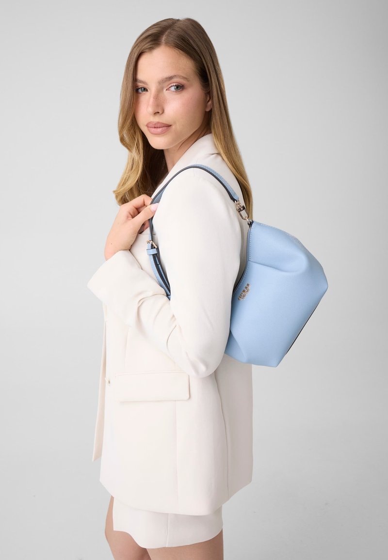 Jeune femme en blazer blanc portant un sac à bandoulière bleu clair avec une forme structurée et des ferrures argentées.