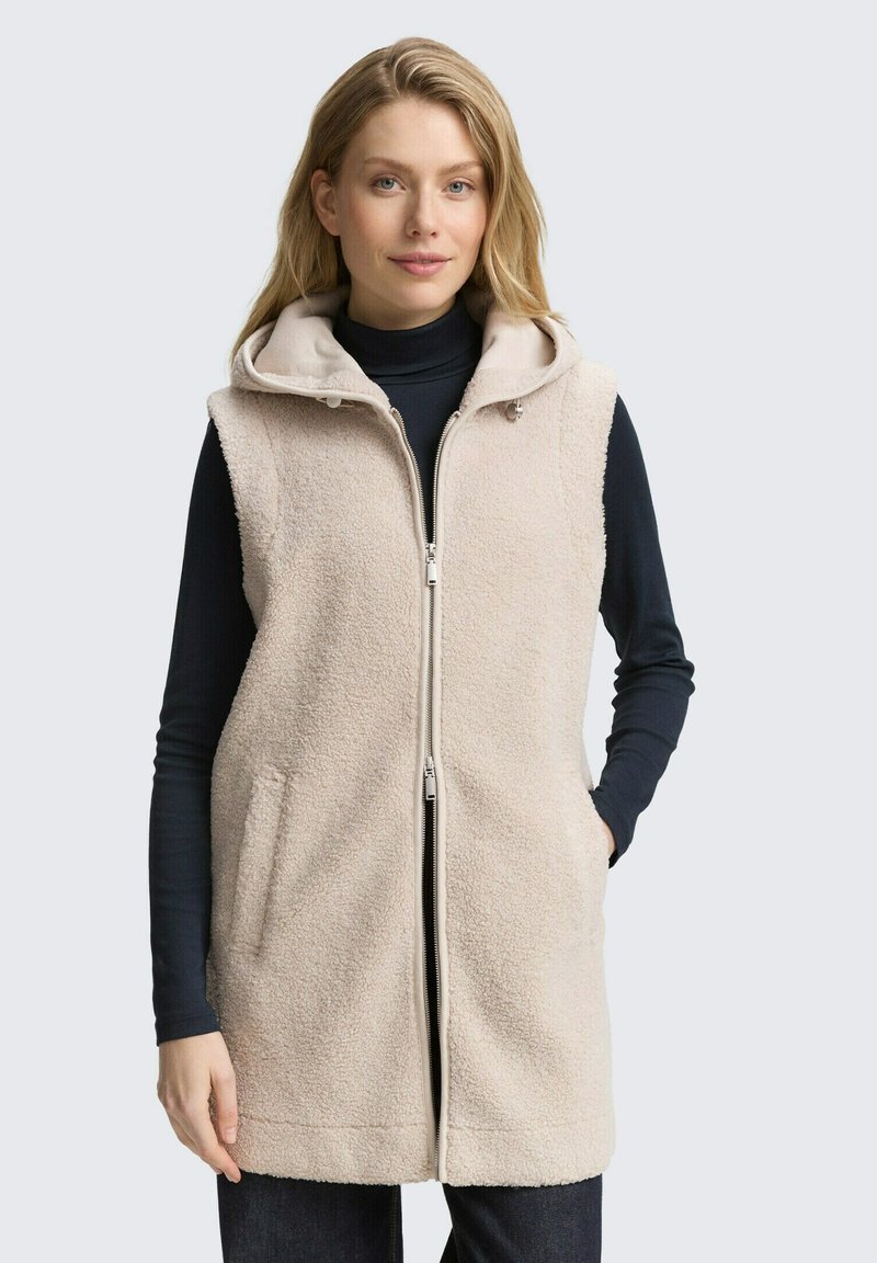Beige Imitationsfellweste mit Reißverschluss vorne, Kapuze und zwei seitlichen Taschen. Das Kleidungsstück zeichnet sich durch eine weiche Textur und einen entspannten Schnitt aus.