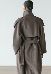 Donna con capelli raccolti in uno chignon che indossa un lungo cappotto marrone di lana con cintura, colletto alto, mostrine sulle spalle e falda posteriore, in piedi davanti a uno sfondo neutro.