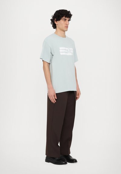 Ανοιχτό μπλε βαμβακερό T-shirt με κοντά μανίκια, που διαθέτει λευκή ριγωτή γραφική απεικόνιση και τον αριθμό 6 μπροστά, φορεμένο με καφέ παντελόνι.