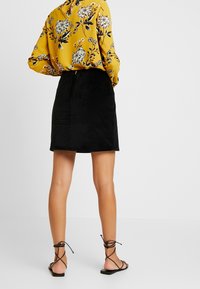 Falda de pana negra con forma recta, combinada con una blusa amarilla de estampado floral y sandalias negras de tiras. La textura es visible.