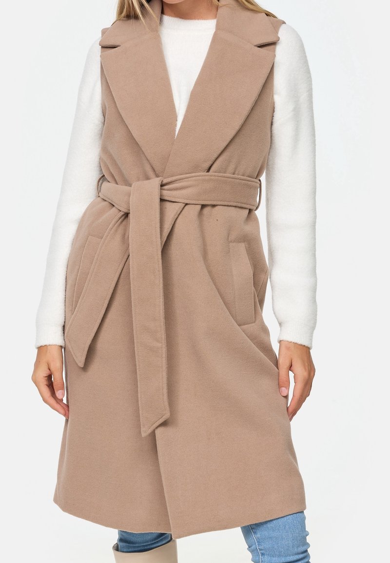 Veste beige sans manches en matériau doux, avec ceinture à la taille, deux poches avant et un col.