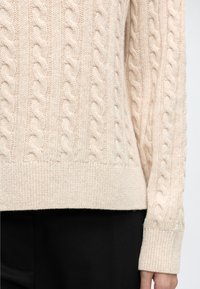 Pull torsadé couleur crème à manches longues porté sur un pantalon noir, montrant le ventre et le bras le long du corps.