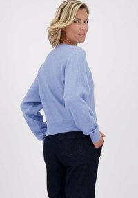 Vrouw met blond haar in een lichtblauwe trui en donkere jeans, poserend met handen in de zakken, kijkt over haar schouder.