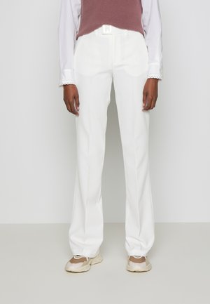 Person trägt weiße gerade geschnittene Hose mit weißem Gürtel, weißes Hemd, braunes ärmelloses Oberteil und beige Sneakers, steht auf einem grauen Boden.