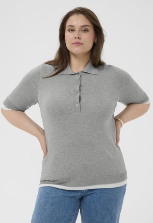 Ung kvinde med brunt hår iført en grå, knappet kortærmet polo-shirt og blå jeans, stående med hænderne på hofterne.