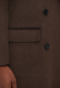 Barna gyapjú kabát, amely vállra szabott fazonú, egysoros gombos záródással és oldalzsebbel rendelkezik. A textúrája puha és sima.