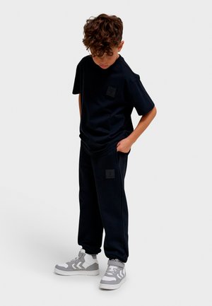 Sort t-shirt med korte ærmer og en firkantet logo-patch, kombineret med matchende joggers med sidelommer og logo-patches. Fuldført med grå sneakers.