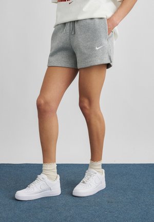 Persoană purtând pantaloni scurți Nike gri, adidași albi și șosete crem stând pe un covor albastru cu mâinile în buzunare.