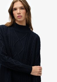 Jersey de punto azul marino extragrande con cuello alto, patrones texturizados de trenza y puños acanalados, hecho de un hilo suave.