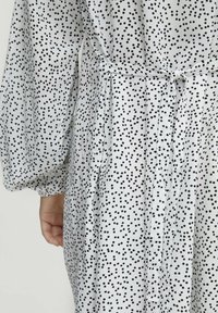 Weißes Kleid aus Stoff mit schwarzen Pünktchen, das lange Puffärmel, eine taillierte Taille mit Bändern und Raffungen am Rücken aufweist.