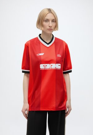 Donna con capelli biondi corti che indossa una maglia sportiva rossa larga con colletto e polsini neri e bianchi, in piedi su sfondo bianco.