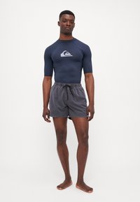 Tengerész navy színű rövidujjú rash guard és szürke fürdőnadrág, amely rugalmas derékkal és fekete zsinórral rendelkezik; a modell mezítláb áll.
