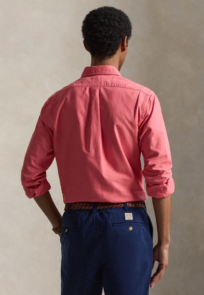 Muž s kudrnatými vlasy stojí zády, má na sobě korálově dlouhý rukáv s vyhrnutými rukávy a tmavě modré kalhoty s pleteným páskem.