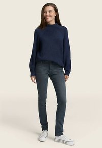 Tamsiai mėlynas megztinis su iškiliais raštais ir aukšta apykakle, derinamas su pilkomis lieknėjančiomis džinsais ir baltais sportiniais bateliais. Audinys atrodo stambus ir minkštas.