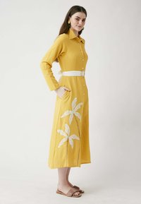 Keltainen pitkähihainen mekko, jossa on valkoinen vyö, kukkakirjonta, napit edessä ja sivutaskut. Mallilla on päällään ruskeat sandaalit.