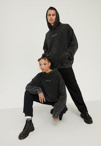 Dva modela nose crne hoodice, jedan stoji s rukama u džepovima, dok drugi čuči noseći crne čizme i hlače, na bijeloj pozadini.