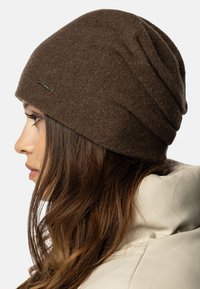 Brun strikhat med en blød tekstur og et slouchy design, der har et lille logomærke på siden. Profileret udsigt med langt hår synligt.