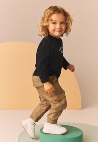 Barns outfit med en svart långärmad tröja och khakifärgade cargobyxor. Vita sneakers med texturerade sulor. Står på en grön piedestal.