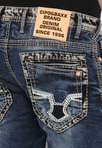 Blaue Denim-Jeans mit einem auffälligen Design der Gesäßtasche, bestickten Akzenten, einem markierten Lederpatch und sichtbaren Nähdetails.