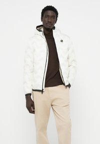 Witte gewatteerde jacket met een capuchon, voorzien van een chevronpatroon en zwarte accenten. Draagt over een bruine shirt, in combinatie met beige broek.