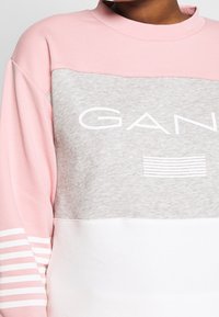 Sweatshirt s blokováním barev, který má růžové, šedé a bílé části. Měkká tkanina, kulatý výstřih a bílá logo s detailními horizontálními pruhy.