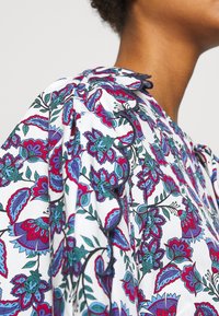 Gros plan sur une épaule portant un chemisier blanc avec un motif floral coloré et des bords festonnés bordés de fil foncé.