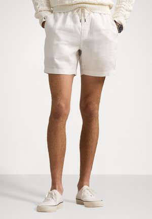 Homme portant un pull en maille crème, un short à cordon de serrage blanc cassé, des baskets blanches, debout les mains dans les poches sur un fond uni.