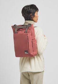 Rode nylon rugzak met grijze banden, voorzak met rits en logo-details. Heeft een rechthoekige vorm en een minimalistisch ontwerp.