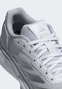 Biele športové topánky s textúrovaným sieťovinovým zvrškom, hladkými koženými doplnkami a šnúrkami. Majú výrazné logo značky a polstrovaný lem.