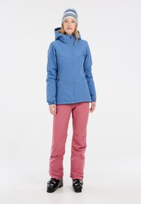 Blaue Softshelljacke mit Kapuze, pinke Hose und schwarze Skischuhe. Die Jacke verfügt über einen Reißverschluss vorne und geometrische Nähte.