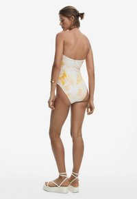 Maillot de bain une pièce blanc avec un motif aquarelle jaune, cuisses hautes et sans bretelles. Porté avec des sandales à plateforme blanches et des bijoux discrets.