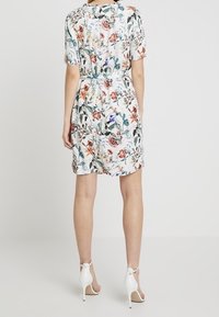 Mujer vistiendo un vestido blanco de manga corta y longitud a la rodilla con estampado floral multicolor y sandalias blancas de tacón alto con pulsera al tobillo, vista desde atrás.