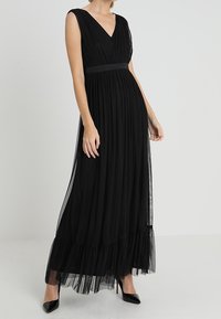 Černé maxi šaty s hlubokým V výstřihem, plisovaným korpusem a vrstveným tylovým lemem. Mají přiléhavý pas a bezrukávový design.